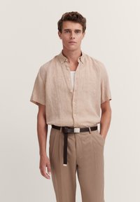 Kurzärmliges, hellbeiges Leinentat mit spitzem Kragen und Brusttasche, kombiniert mit hoch taillierten, hellbraunen Hosen und einem dunklen Gürtel.