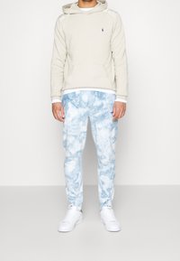 Sweat à capuche beige avec une poche avant et un logo, associé à un pantalon de jogging tie-dye bleu. Des baskets blanches complètent la tenue.