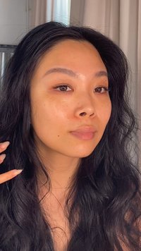 Gros plan sur un visage avec de longs cheveux noirs ondulés. La peau est lisse avec une éclat naturel. Les traits incluent des yeux marron et une couleur de lèvres légère et subtile.