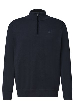 Homme aux cheveux courts, bouclés et foncés, portant un pull bleu marine à fermeture éclair quart sur une chemise blanche et un jean bleu, debout devant un fond uni.