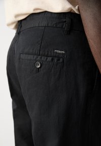 Black pants made of a textured fabric, featuring a rear pocket with a button closure and a woven label on the waistband.