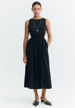 Frau trägt ärmelloses schwarzes Midikleid mit Ausschnitten an den Seiten, lange Halskette mit Anhänger und grüne spitze Pumps mit niedrigem Absatz.