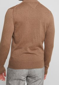 Mand iført en brun langærmet sweater og grå teksturerede bukser, vist fra ryggen mod en ensfarvet baggrund.
