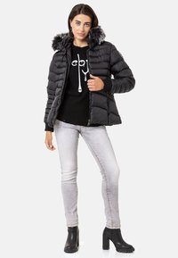 Schwarze Daunenjacke mit einer Kapuze aus Kunstfell, Reißverschluss, Seitentaschen und Steppung. Getragen über einem schwarzen Pullover und hellen Jeans.