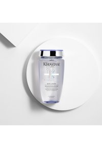 Flacon de shampooing hydratant et illuminant Kérastase Blond Absolu Bain Lumière sur une plateforme circulaire blanche, fond clair.
