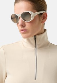 Białe okulary przeciwsłoneczne z okrągłymi oprawkami i przyciemnianymi soczewkami, zestawione z beżowym topem zapinanym na zamek z wysokim kołnierzem, o dopasowanym kroju i prostych szwach.