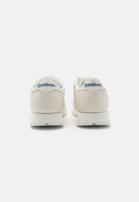 Šviesiai smėlio spalvos Reebok sportbačiai su lygaus odos viršumi, balta padais ir mėlynu logotipu ant kulno, su paminkštintu apykakle ir tekstūruotu padu.