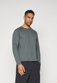 Chemise grise à manches longues côtelée avec un col rond et des manches raglan, portée avec un pantalon sombre. Le tissu a une finition texturée.