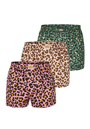 Shirtless man with curly hair and beard standing with arms crossed, wearing pink leopard print boxer shorts against a plain background.