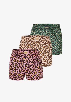 Hemdloser Mann mit lockigem Haar und Bart, der mit verschränkten Armen steht und pinke Leoparden-Boxershorts vor schlichtem Hintergrund trägt.