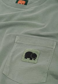 Světle zelené bavlněné tričko s přední kapsou, na které je černý sloní logo na zeleném látkovém štítku. Měkká struktura, neformální design.