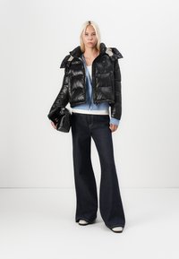 Schwarze Pufferjacke mit glänzender Oberfläche, cropped Design, großer Kragen. Kombiniert mit einem hellblauen Cardigan, dunklen weiten Jeans und einer schwarzen Clutch.