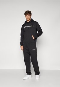Ensemble en polaire comprenant un sweat à capuche noir et un pantalon, orné d'un grand logo "Champion" blanc, avec une taille élastique et des poches, associé à des baskets blanches.