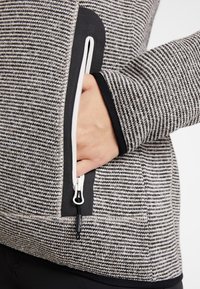 Veste en maille noire et blanche avec une poche zippée. Présente un motif texturé, une bordure noire contrastante et une fermeture éclair avec languette de tirage.