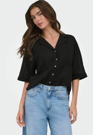 Jeune femme aux longs cheveux bruns portant une chemise noire à manches courtes avec boutons et un jean bleu clair taille haute, posant devant un fond blanc.
