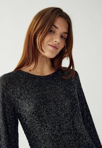 Tânără cu păr drept, castaniu, purtând un top negru cu paiete strălucitoare, zâmbind ușor pe un fundal deschis.