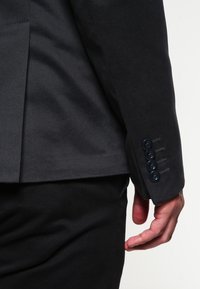 Zbliżenie na osobę w czarnej marynarce z czterema guzikami na mankiecie, której ręka swobodnie spoczywa przy boku.