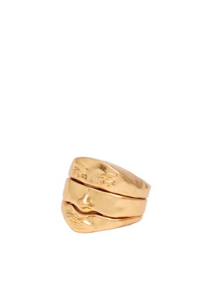 Anillo de metal color oro en forma de rostro humano partido con ojos, nariz y labios detallados sobre fondo blanco.