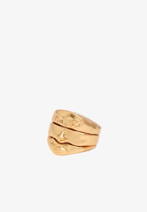 Anillo de metal color oro en forma de rostro humano partido con ojos, nariz y labios detallados sobre fondo blanco.