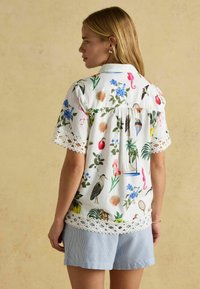 Floral-patterned white blouse with short sleeves and lace trim, featuring colorful birds and plants, paired with light blue striped shorts.