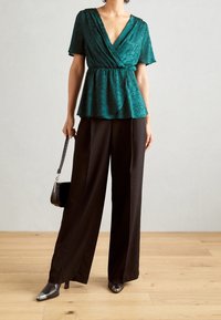 Blusa avvolgente teal con scollo a V e maniche corte, abbinata a pantaloni neri a gamba larga. Presenta un tessuto strutturato e un dettaglio con vita legata.