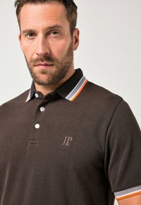 Polo marrón con diseño de cuello, tres botones y franjas de acento en naranja y blanco contrastantes en el cuello, con un logo en el pecho.