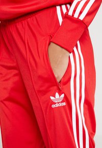 Röda träningsbyxor i slät tyg med vitt tre-stripes mönster på sidorna och en vit logotype. Har en sidoficka.