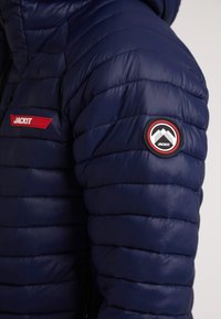 Marineblå pufferjakke med kviltet design, der har et rødt logo-patch og et rundt sort mærke på skulderen. Glat, blankt stof.