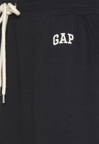 Primo piano di pantaloni sportivi neri in cotone con cordino bianco e logo GAP ricamato bianco nella zona della tasca anteriore.