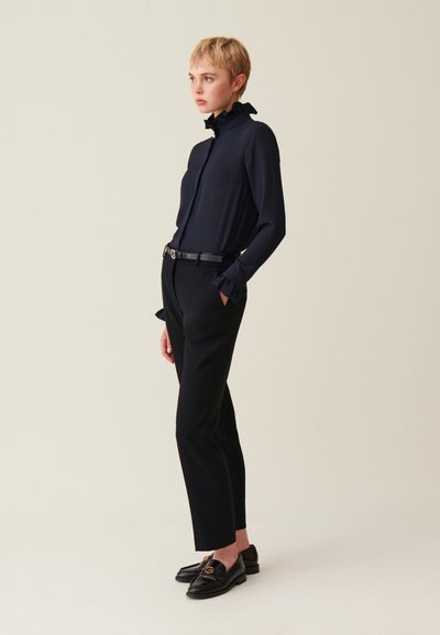 Blusa blu navy con colletto a volants, taschino frontale e maniche lunghe; abbinata a pantaloni neri su misura e scarpe nere con dettagli dorati.