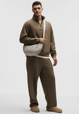 Mann trägt braune Sweatjacke mit Reißverschluss und Hose, dazu eine beige Umhängetasche und passende Slip-On-Schuhe vor einfarbigem Hintergrund.