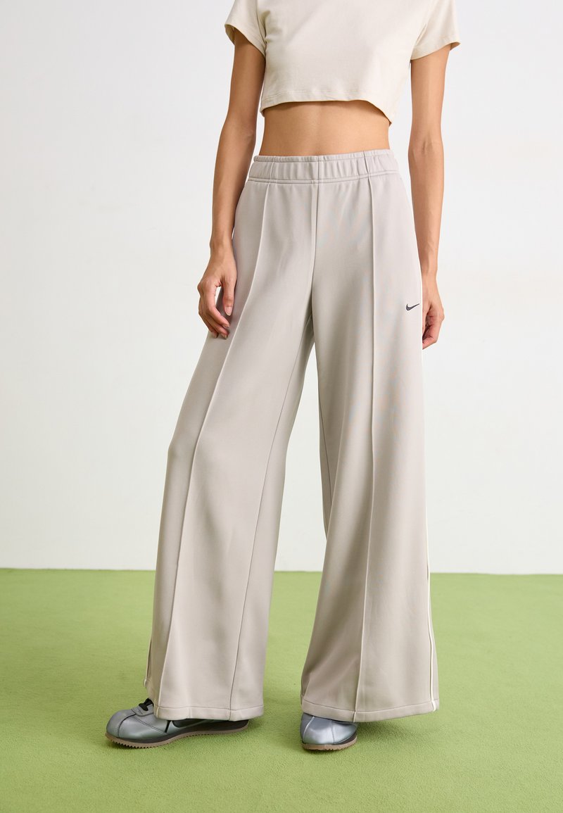 Pantalons de survêtement Nike gris clair avec une coupe ample, taille élastique et bandes latérales, associés à des chaussures grises sur un sol vert.