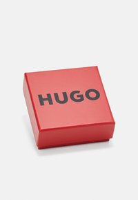 Scatola regalo rettangolare rossa con finitura lucida, con la parola "HUGO" in carattere nero in grassetto sulla superficie superiore.