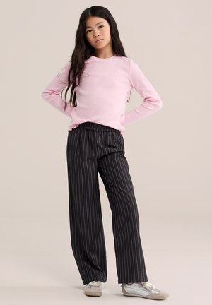 Fille aux longs cheveux foncés, debout les mains sur les hanches, portant un haut rose à manches longues, un pantalon large noir à rayures fines et des baskets blanches.
