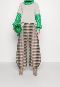 Grüner, gestrickter Pullover mit gestreiften Ärmeln, kombiniert mit weit geschnittenen Hosen in mehrfarbigen Streifen. Die Accessoire ist eine grüne Handtasche.