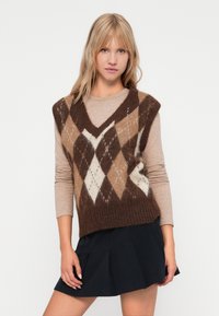 Brun og creme argyle-mønstret strikket vest med V-hals, lagt over en beige langærmet top. Båret med en mørk plisseret nederdel.