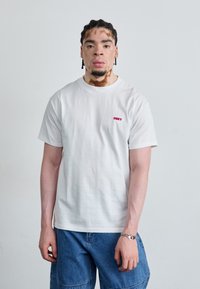 T-shirt branca de algodão com um pequeno logótipo vermelho "OBEY" no peito, acompanhada de calções de ganga azuis. Textura suave, design casual.