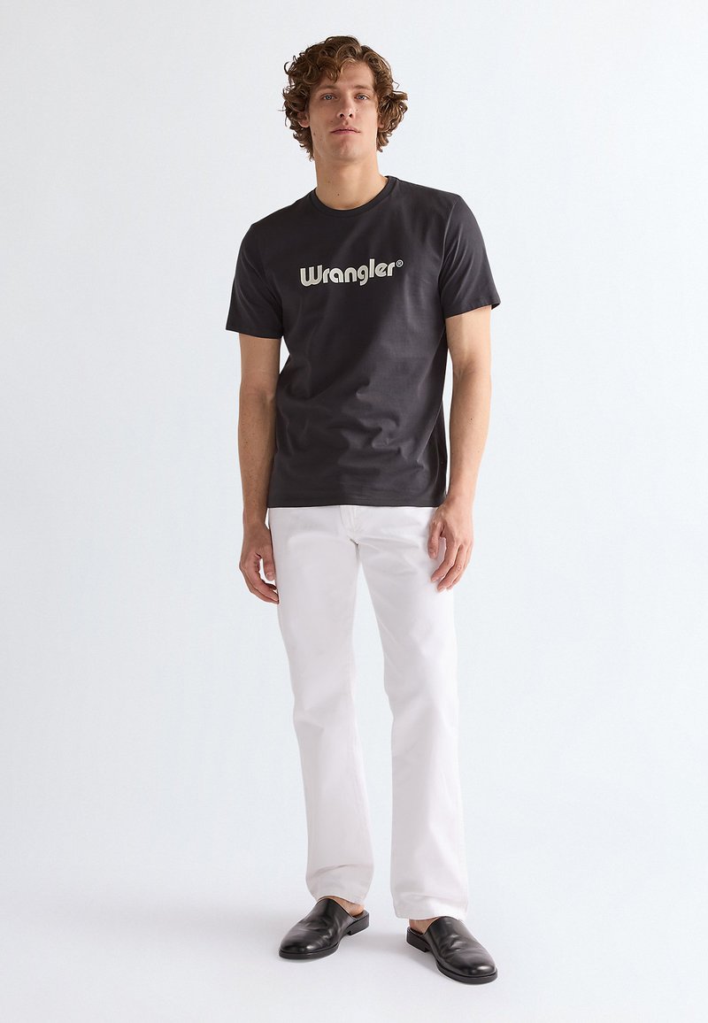 Mladý muž s kučeravými vlasmi, ktorý má na sebe čierne tričko Wrangler, biele nohavice a čierne nazúvacie topánky, stojí pred jednofarebným bielym pozadím.