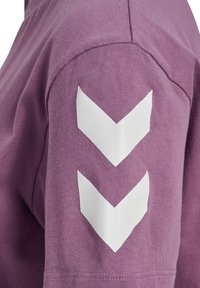 Fioletowa koszula z krótkim rękawem z trzema białymi wzorami w kształcie litery V na lewym rękawie. Materiał z mieszanki bawełny o gładkiej strukturze.