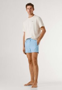 Ung mand står barfodet iført en hvid kortærmet skjorte og lyseblå shorts med snøre foran mod en ensfarvet baggrund.