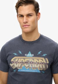 Camiseta de algodón gris con un gran y audaz gráfico de "SUPERDRY" en dorado y azul, situado sobre un diseño de explosión estelar con acentos de relámpago.
