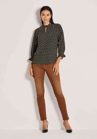 Schwarze gemusterte Bluse mit Schlüsselloch-Ausschnitt und Glockenärmeln, kombiniert mit braunen Slim-Fit-Jeans und schwarzen High Heels. Insgesamt glatte Textur.