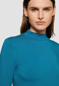 Turquoise lange mouw top met een hoge hals. Zachte textuur, aansluitend ontwerp en minimalistische naden, die een hedendaagse stijl uitstralen.