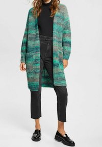 Długość do kolan cardigan w zielonym odcieniu z dzianiny o gradientowej teksturze, z dwoma przednimi kieszeniami i obniżonymi ramionami. Nosi się go na czarnej bluzce i spodniach.