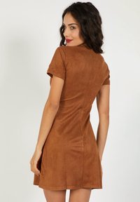 Vestido corto de ante marrón con mangas cortas, cuerpo ajustado y falda acampanada, que presenta una textura suave y detalles de costura mínimos.