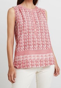Blusa senza maniche rossa e bianca con motivo a diamante e geometrico, dettagli plissettati e scollatura rotonda. Tessuto leggero.