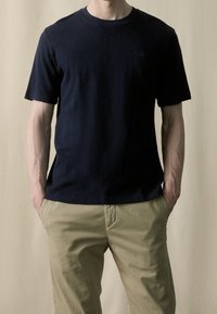 Námořnické modré bavlněné tričko s krátkým rukávem a kulatým výstřihem, v kombinaci se světle béžovými kalhotami. Na hrudi má malé logo.