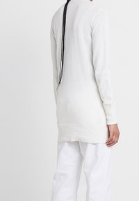Suéter de punto blanco de manga larga con dobladillo acanalado y detalle de costura en la parte posterior, llevado sobre pantalones blancos, con corte recto y textura suave.