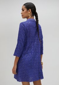 Lila klänning med trekvartslånga ärmar, prydd med ett diamantsmönster och metalliska detaljer. Lätt tyg, med krage och avslappnad passform.