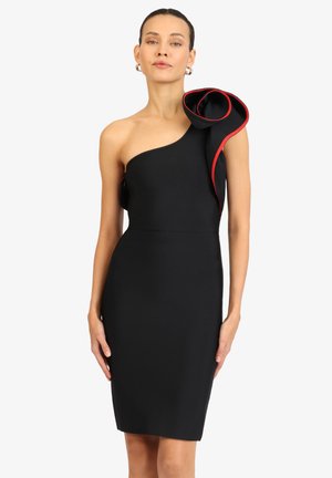 Frau trägt ein schwarzes Ein-Schulter-Kleid mit einem großen, roten Rüschendetail an der Schulter, steht vor einem weißen Hintergrund.